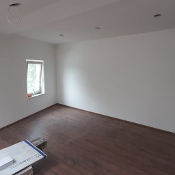 Art-House Jarosław Zaborowski - Pusta, jasna przestrzeń z brązową podłogą imitującą drewno, widoczne niedokończone otwory na oświetlenie w suficie oraz kartony z płytkami ceramicznymi przy ścianie, sugerujące prace wykończeniowe.