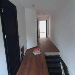 Art-House Jarosław Zaborowski - Korytarz w trakcie remontu z nową, brązową podłogą imitującą drewno, czarnymi drzwiami i schodami, widoczne niedokończone punkty oświetleniowe w suficie i instalacja hydrauliczna za drzwiami.