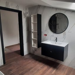 Art-House Jarosław Zaborowski - Nowoczesna łazienka z marmurowymi płytkami na ścianach, drewnopodobną podłogą, okrągłym lustrem, czarną szafką pod umywalką i wysoką szafką wiszącą z otwartymi półkami.
