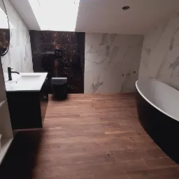 Nowoczesna łazienka na poddaszu z drewnopodobną podłogą, czarną armaturą i marmurowymi płytkami na ścianach; widoczne otwory w suficie na oświetlenie.