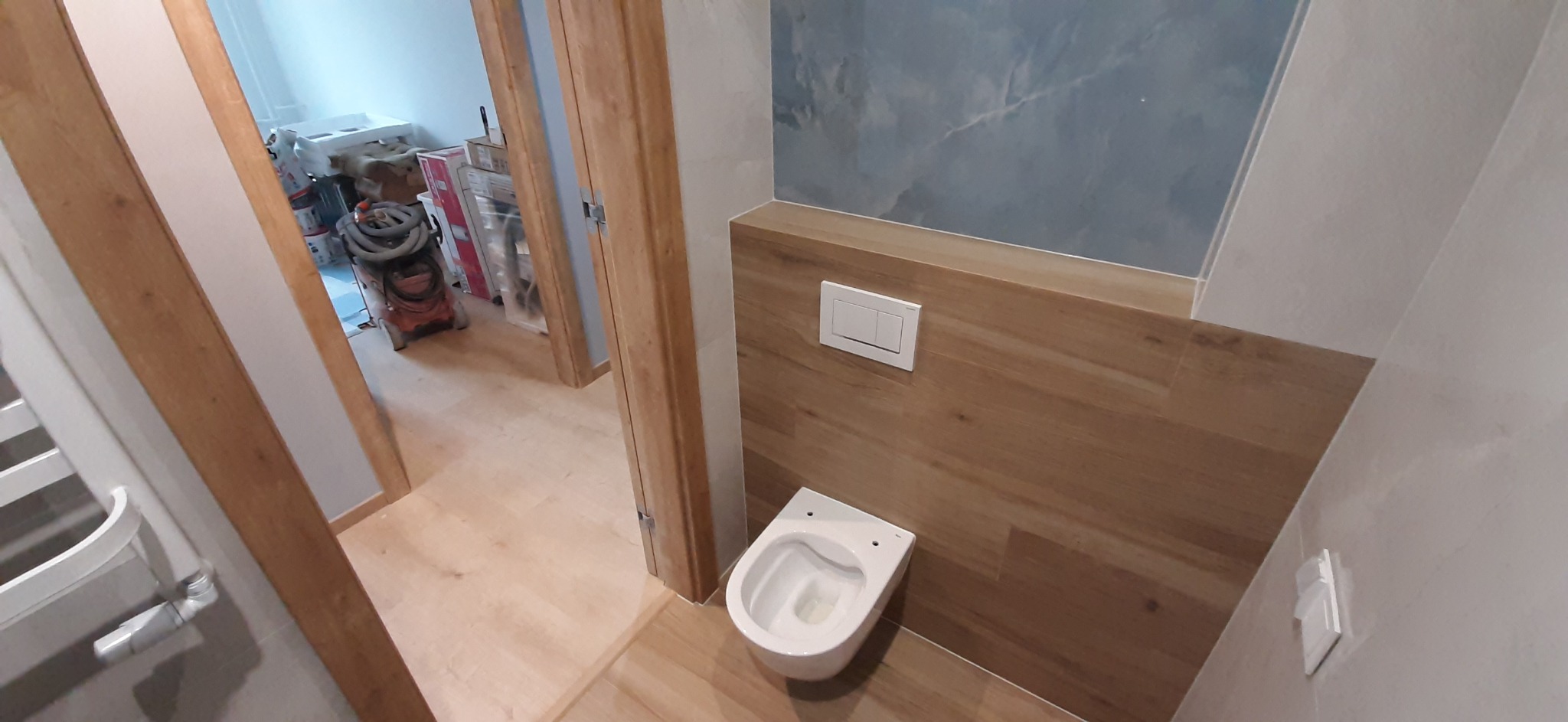 Wnętrze łazienki w trakcie remontu, widoczna podwieszana toaleta, drewnopodobne panele na ścianie i niebieska ściana imitująca marmur, w tle widoczne narzędzia i materiały budowlane.