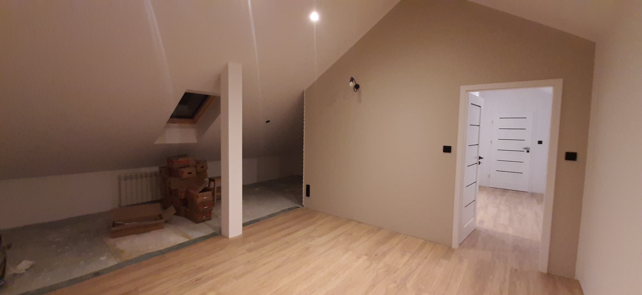 Wnętrze poddasza w Niechodzinie w trakcie wykańczania: jasne panele podłogowe, beżowe ściany, białe drzwi z poziomymi frezami, widoczne kartony i grzejnik w oddzielonej części pomieszczenia, okno...