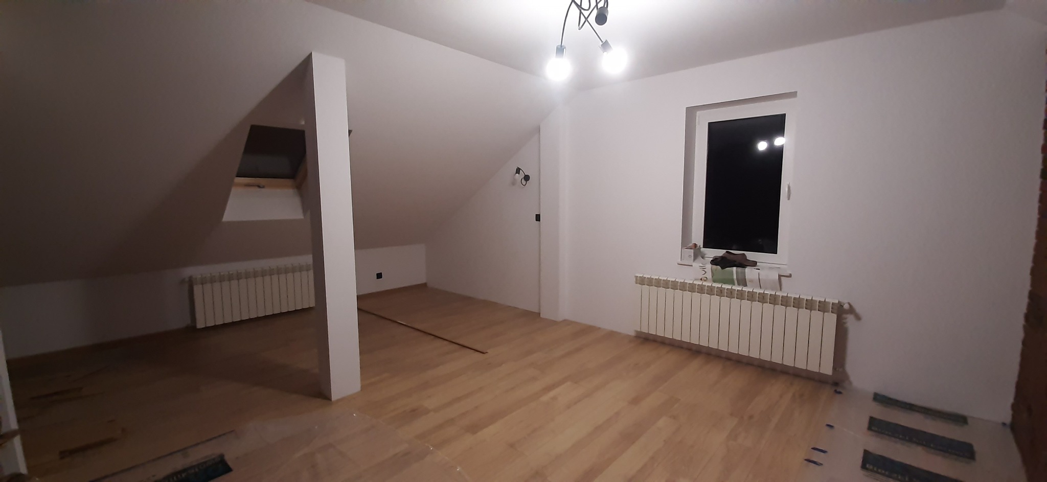 Jasne drewniane panele podłogowe w nowym pokoju na poddaszu z oknem, grzejnikiem, białymi ścianami i sufitem oraz nowoczesną lampą wiszącą.