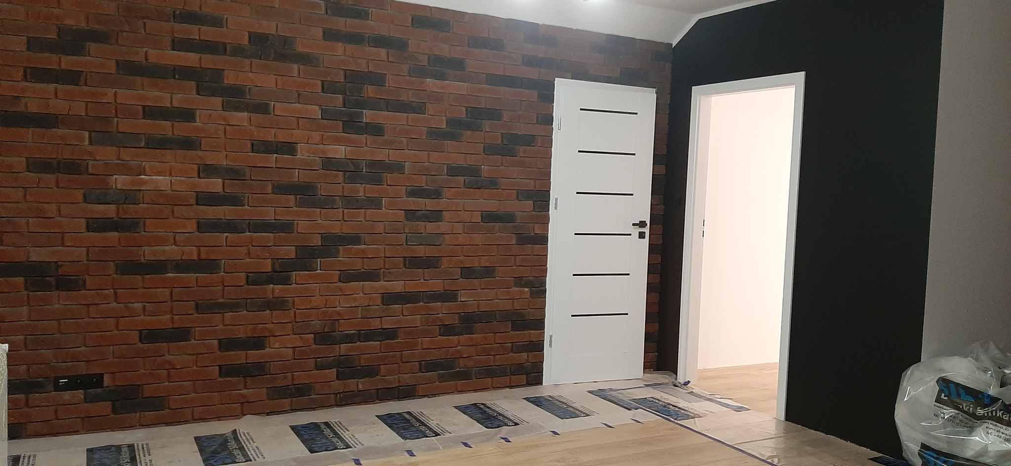 Ściana z cegły klinkierowej w odcieniach czerwieni i czerni, sąsiadująca z białą, nowoczesną ościeżnicą i drzwiami z poziomymi przeszkleniami, na podłodze folia ochronna.