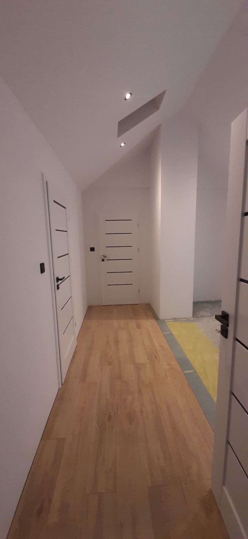 Korytarz w mieszkaniu po remoncie z drewnianą podłogą, dwiema białymi drzwiami z czarnymi klamkami i czarnymi akcentami oraz wbudowanymi punktowymi lampami w białym suficie.