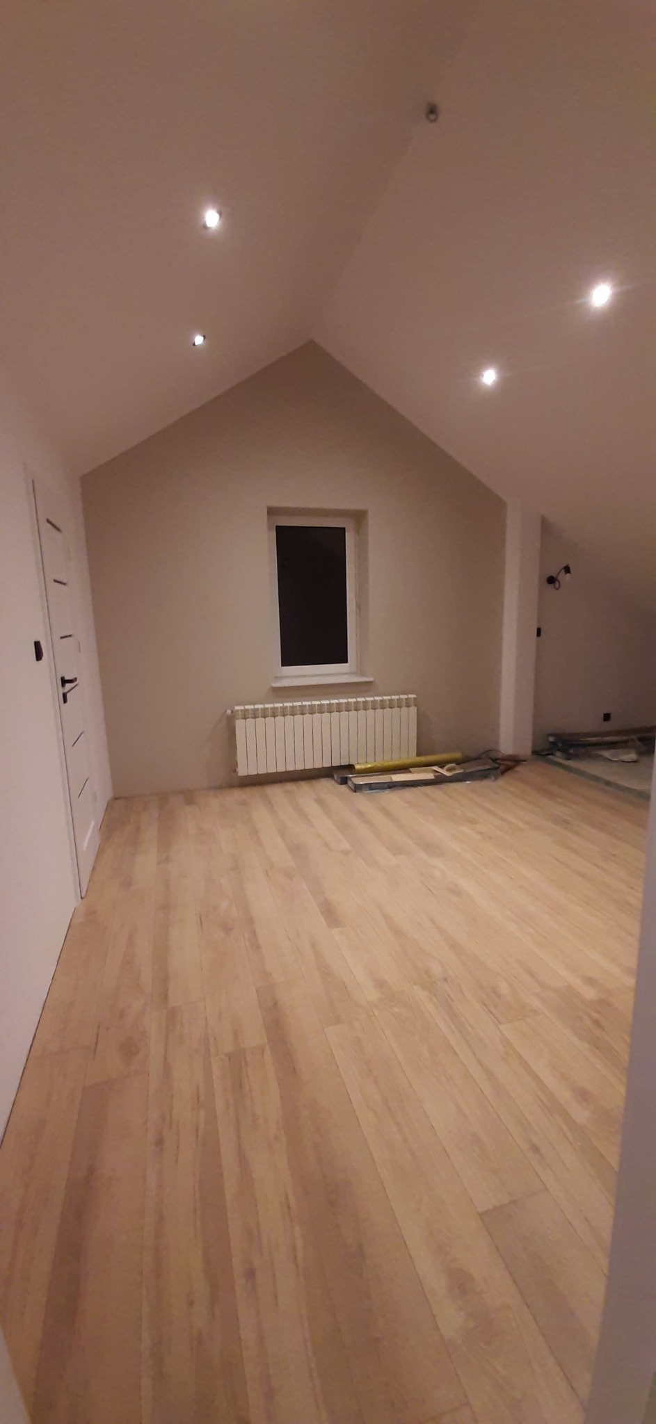 Poddasze po remoncie: jasna podłoga z paneli, białe ściany, okno z grzejnikiem, widoczne narzędzia i poziomica, nowe oświetlenie punktowe.