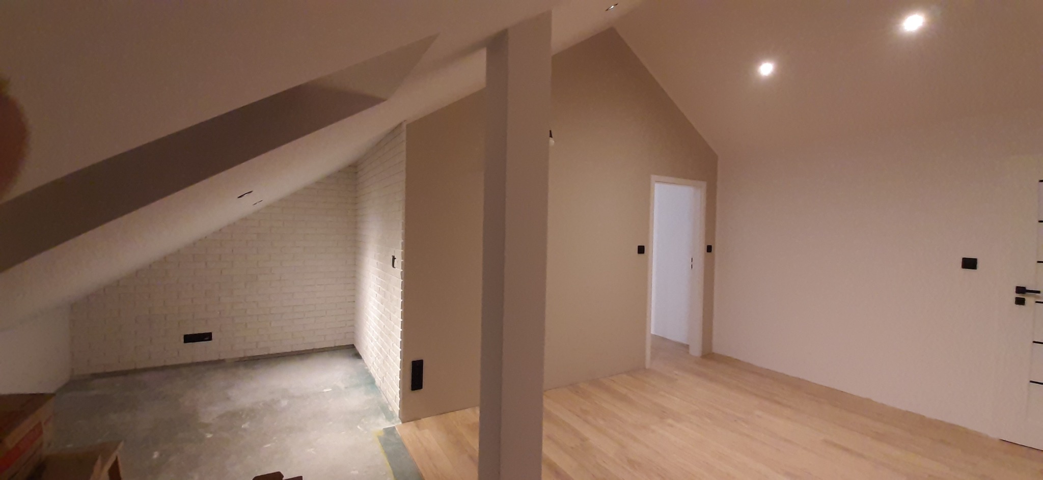 Pokój na poddaszu w trakcie wykańczania: biała cegła na skośnej ścianie, surowa podłoga w jednej części i drewniana podłoga w drugiej, świeżo pomalowane ściany w odcieniach beżu i bieli, widoczne...