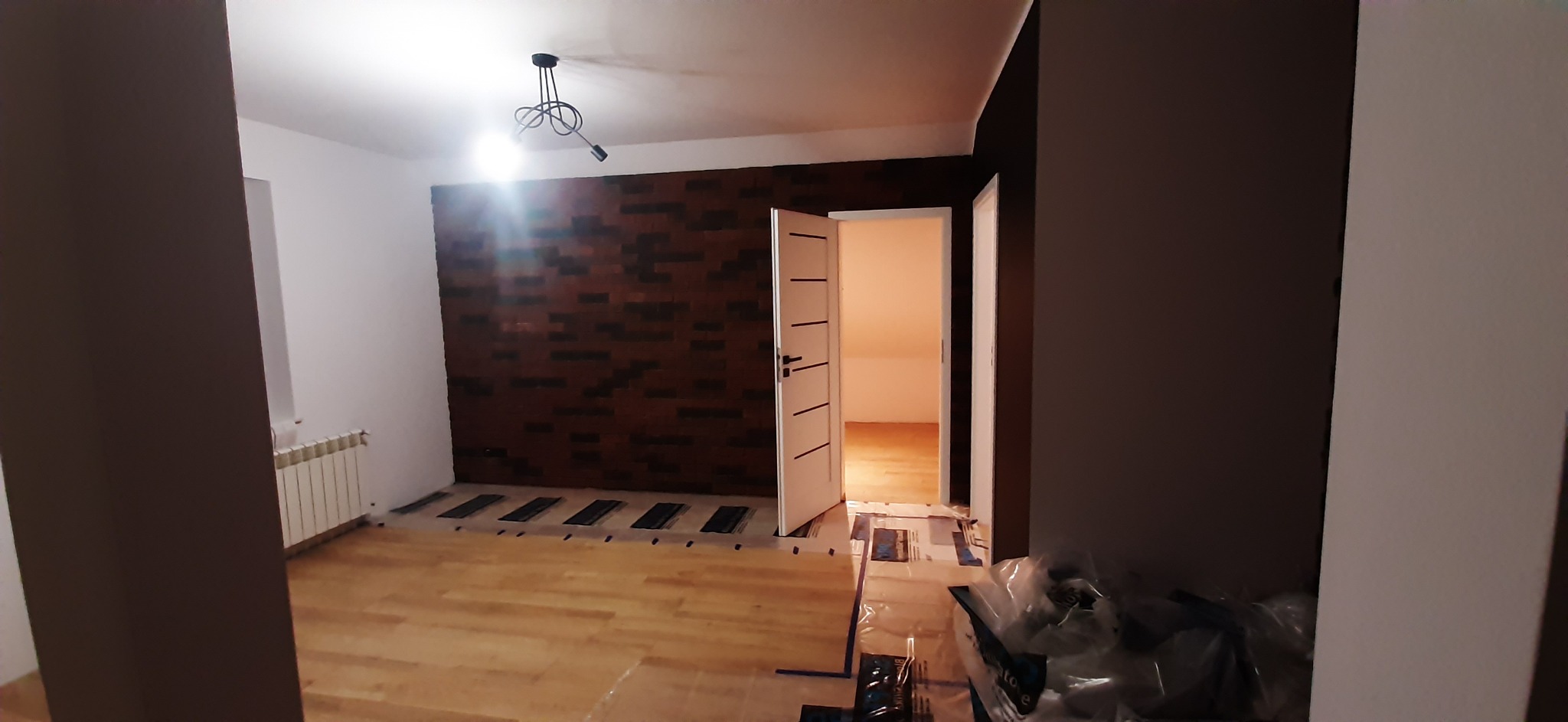 Wnętrze pokoju w trakcie remontu: drewniana podłoga, ściana wyłożona cegłą, białe drzwi, widoczny grzejnik i lampa wisząca.