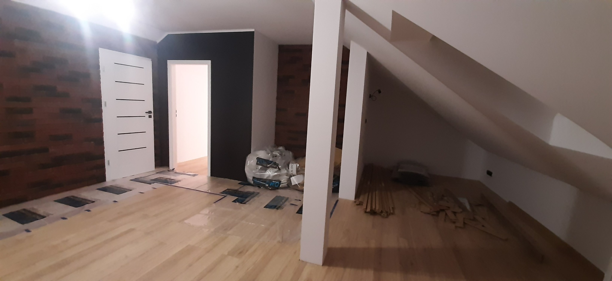 Montaż podłogi drewnianej w pokoju na poddaszu, widoczne przygotowane deski, folia ochronna i fragment ściany z cegły.