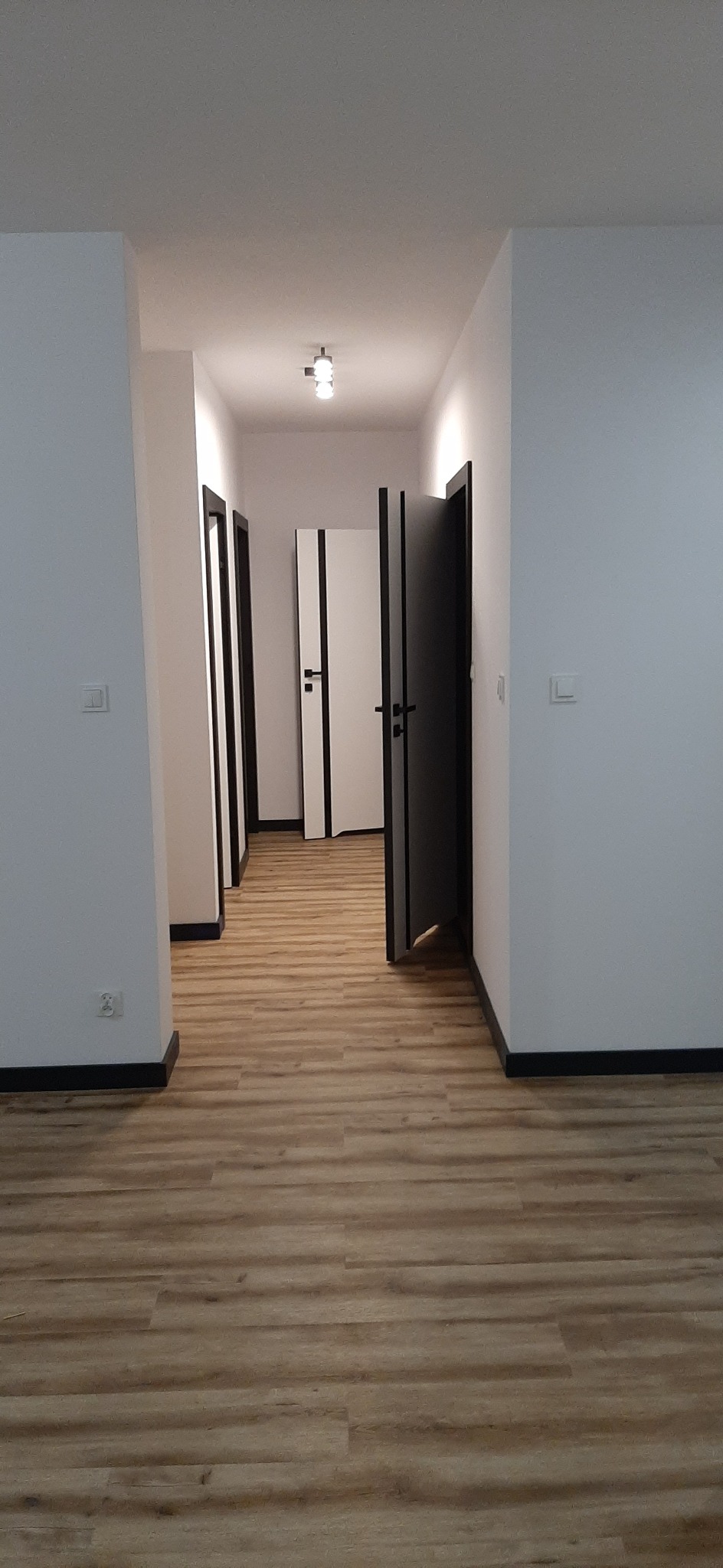 Korytarz w jasnych barwach z panelami podłogowymi imitującymi drewno, białe ściany z czarnymi listwami przypodłogowymi i kilkoma parami drzwi w kolorach białym i czarnym.