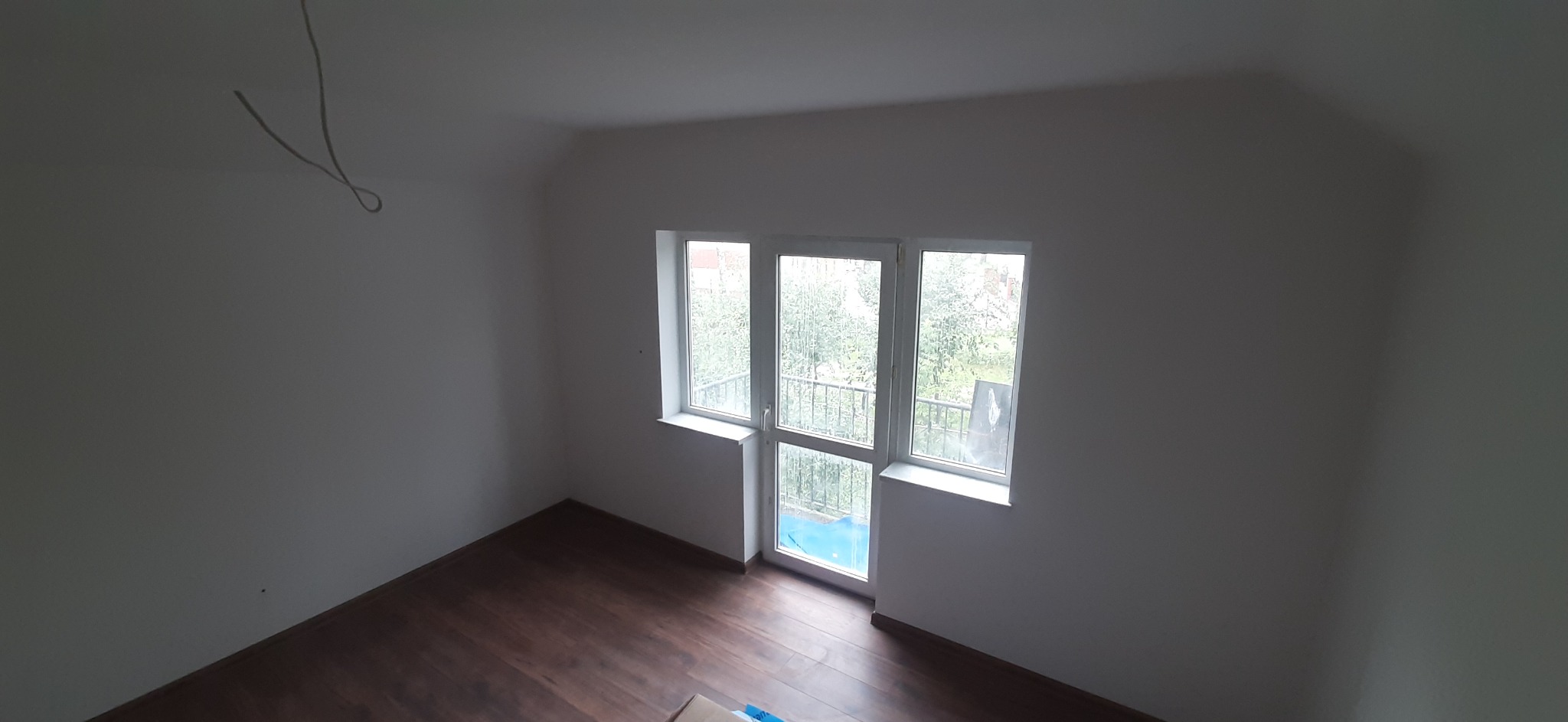 Pusty pokój po remoncie z brązowymi panelami podłogowymi i białymi ścianami, widoczne okno balkonowe z widokiem na zieleń, zwisający kabel elektryczny.