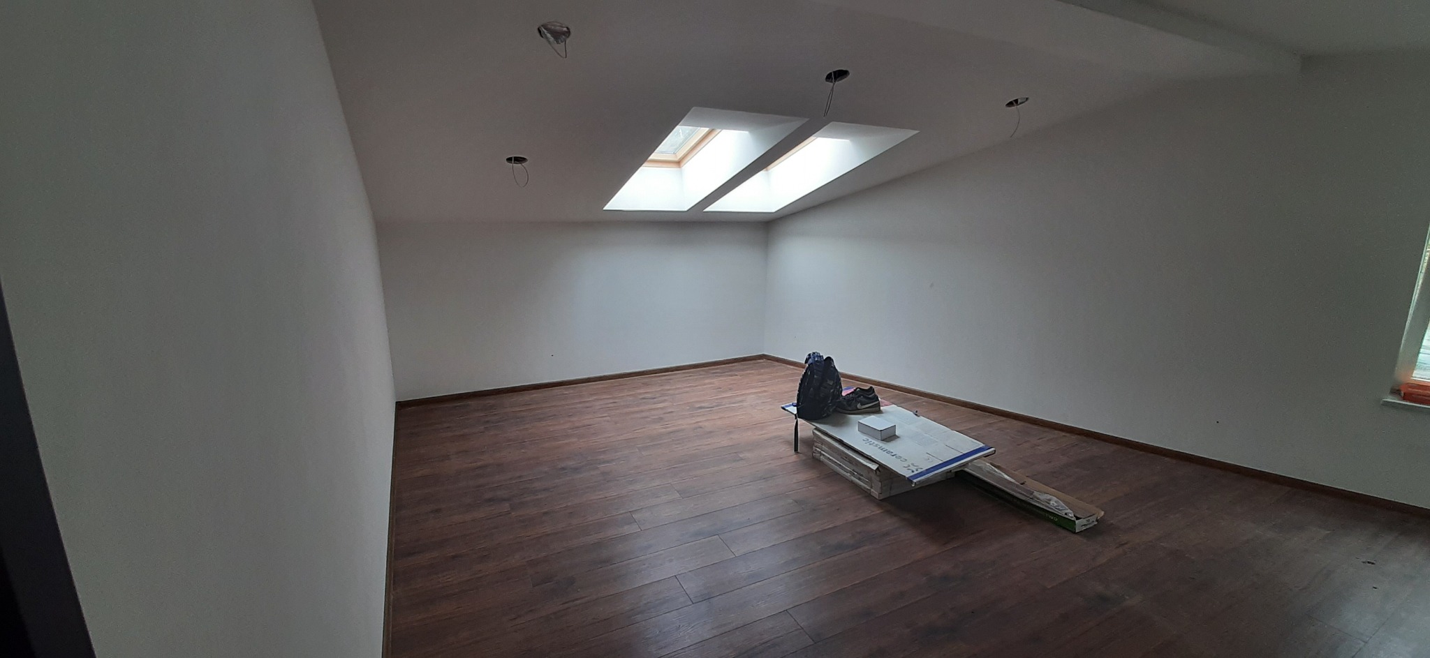 Puste pomieszczenie z drewnianą podłogą, dwoma oknami dachowymi i przygotowanymi otworami na oświetlenie sufitowe, z kartonami i torbą na środku podłogi.