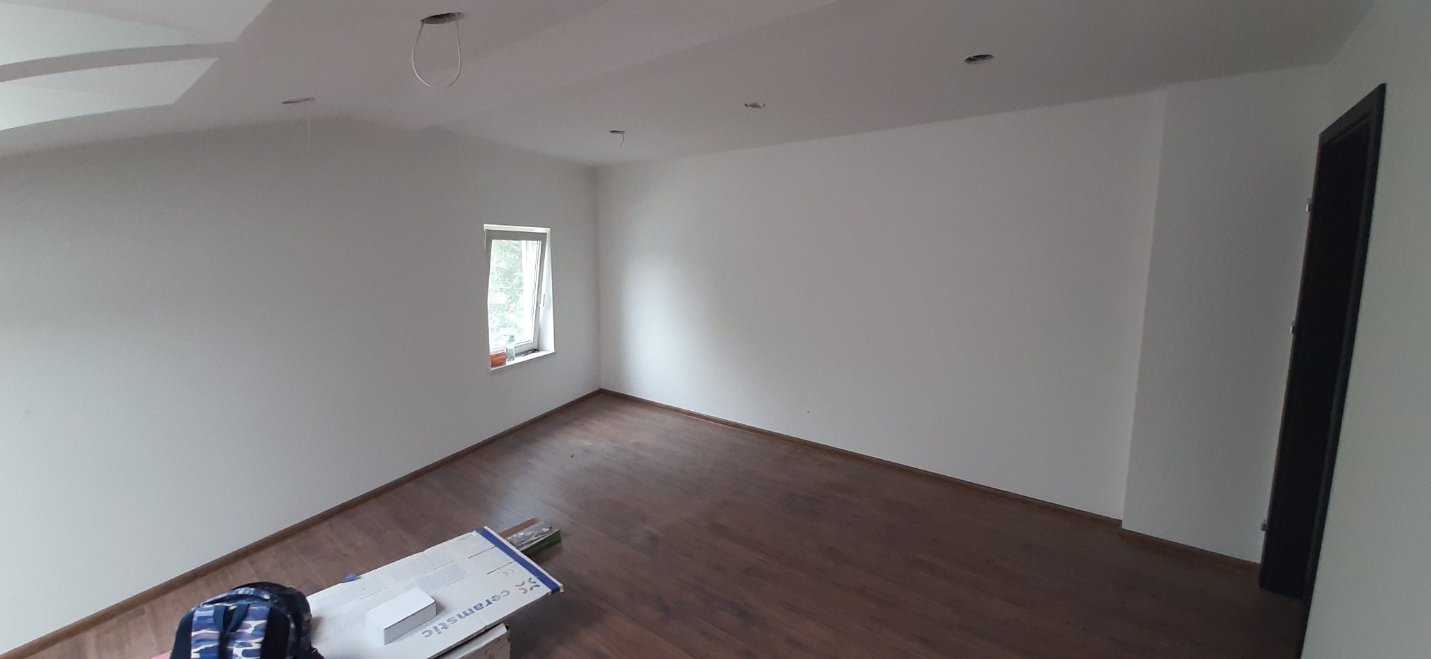 Pusta, jasna przestrzeń z brązową podłogą imitującą drewno, widoczne niedokończone otwory na oświetlenie w suficie oraz kartony z płytkami ceramicznymi przy ścianie, sugerujące prace wykończeniowe.