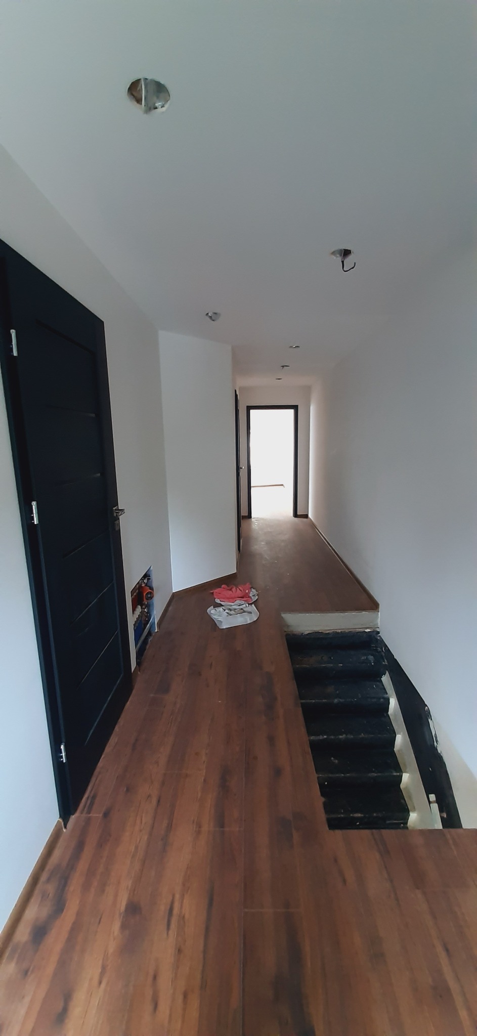 Korytarz w trakcie remontu z nową, brązową podłogą imitującą drewno, czarnymi drzwiami i schodami, widoczne niedokończone punkty oświetleniowe w suficie i instalacja hydrauliczna za drzwiami.