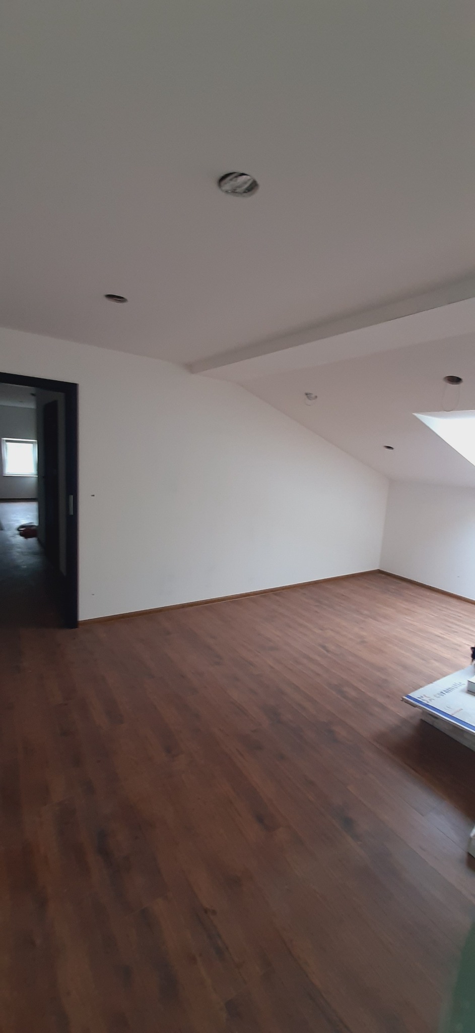 Pokój po remoncie z nową, ciemnobrązową podłogą z paneli, białe ściany i sufit z przygotowanymi otworami na oświetlenie punktowe. Widoczny fragment okna dachowego i drzwi do kolejnego pomieszczenia.