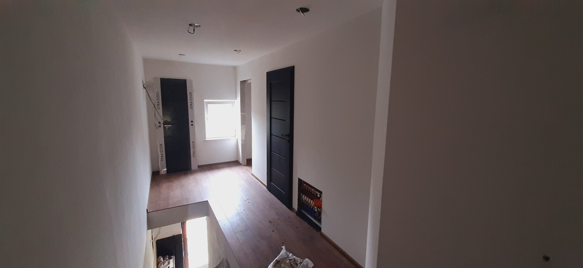 Korytarz w trakcie wykańczania: widoczne dwie czarne drzwi z ościeżnicami marki Stira Door, drewnopodobna podłoga, niezabudowane rury instalacji grzewczej w ścianie, otwory na oświetlenie w suficie.