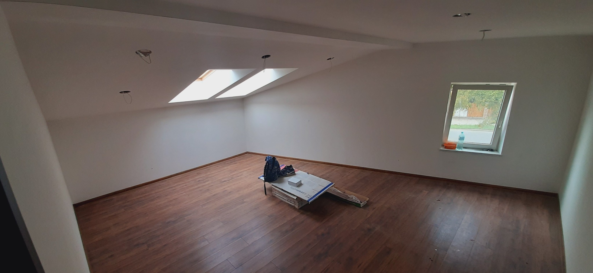 Pokój na poddaszu z drewnianą podłogą, oknem dachowym i standardowym oknem, w trakcie prac wykończeniowych, widoczne niedokończone punkty oświetleniowe na suficie i paczki z materiałami na podłodze.
