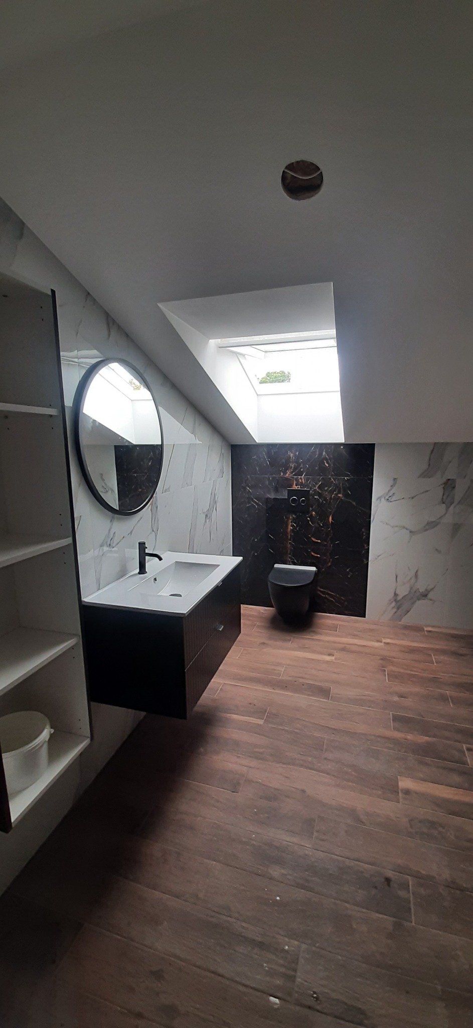 Nowoczesna łazienka na poddaszu z drewnopodobną podłogą, marmurowymi płytkami na ścianach, czarną umywalką wiszącą z okrągłym lustrem, wiszącą toaletą i oknem dachowym.