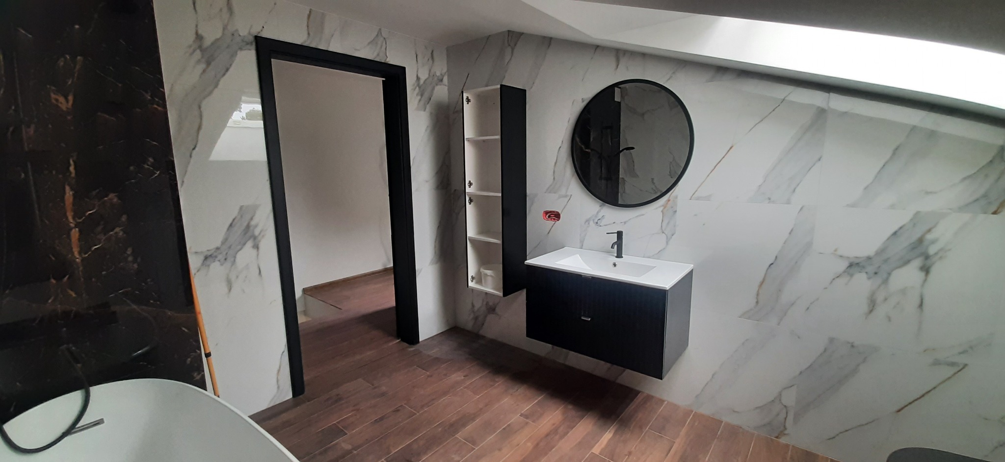 Nowoczesna łazienka z marmurowymi płytkami na ścianach, drewnopodobną podłogą, okrągłym lustrem, czarną szafką pod umywalką i wysoką szafką wiszącą z otwartymi półkami.