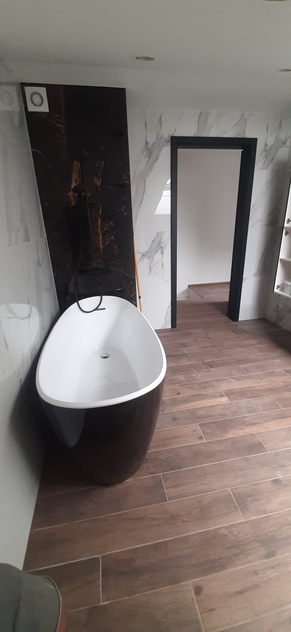 Wolnostojąca wanna w czarno-białej tonacji na tle marmurowych ścian i drewnopodobnej podłogi z płytek, z czarną armaturą i ramą drzwiową.