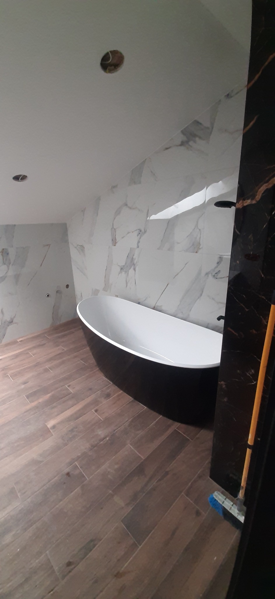 Nowoczesna łazienka w trakcie wykańczania: wolnostojąca wanna w kolorze czarnym, marmurowe płytki na ścianach, drewnopodobna podłoga, widoczne niedokończone otwory na oświetlenie w suficie.