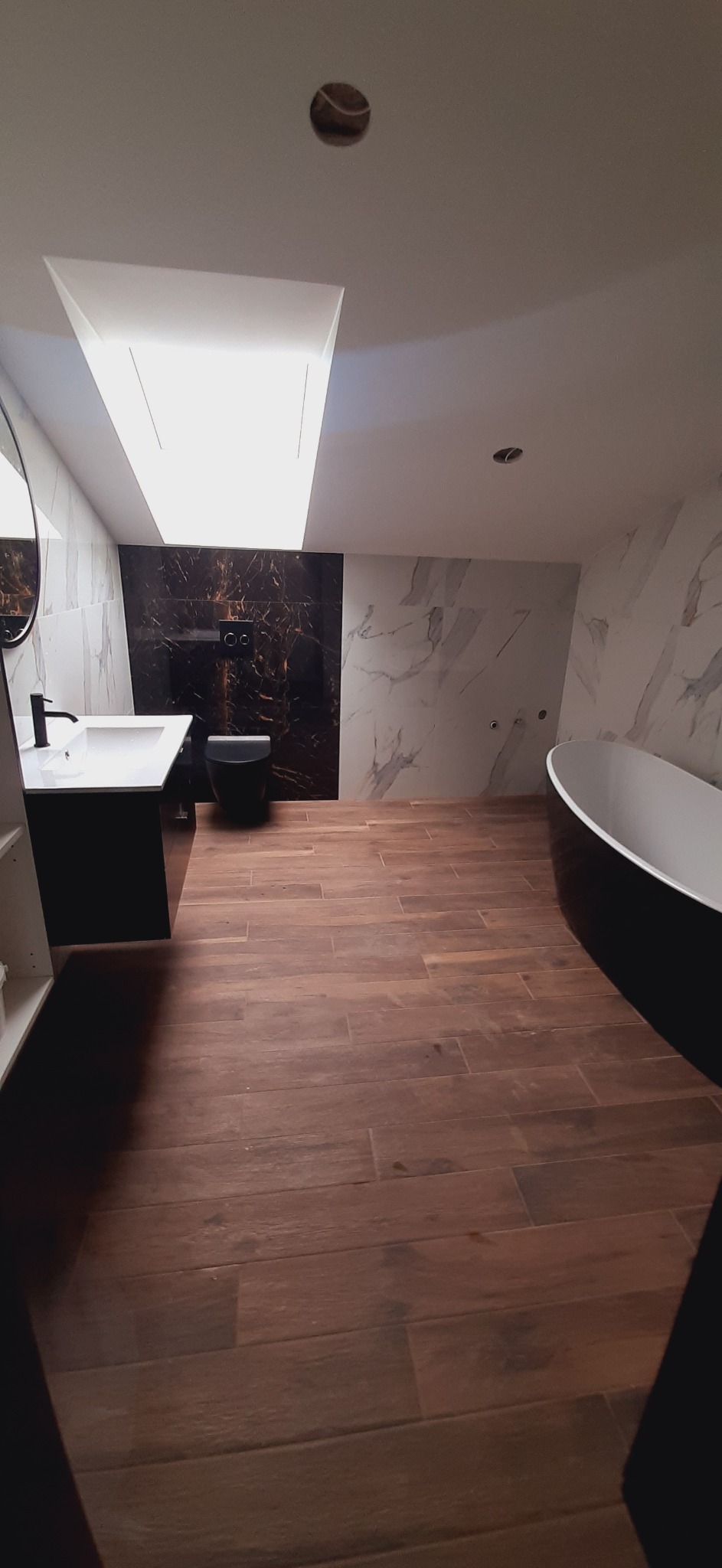 Nowoczesna łazienka na poddaszu z drewnopodobną podłogą, czarną armaturą i marmurowymi płytkami na ścianach; widoczne otwory w suficie na oświetlenie.