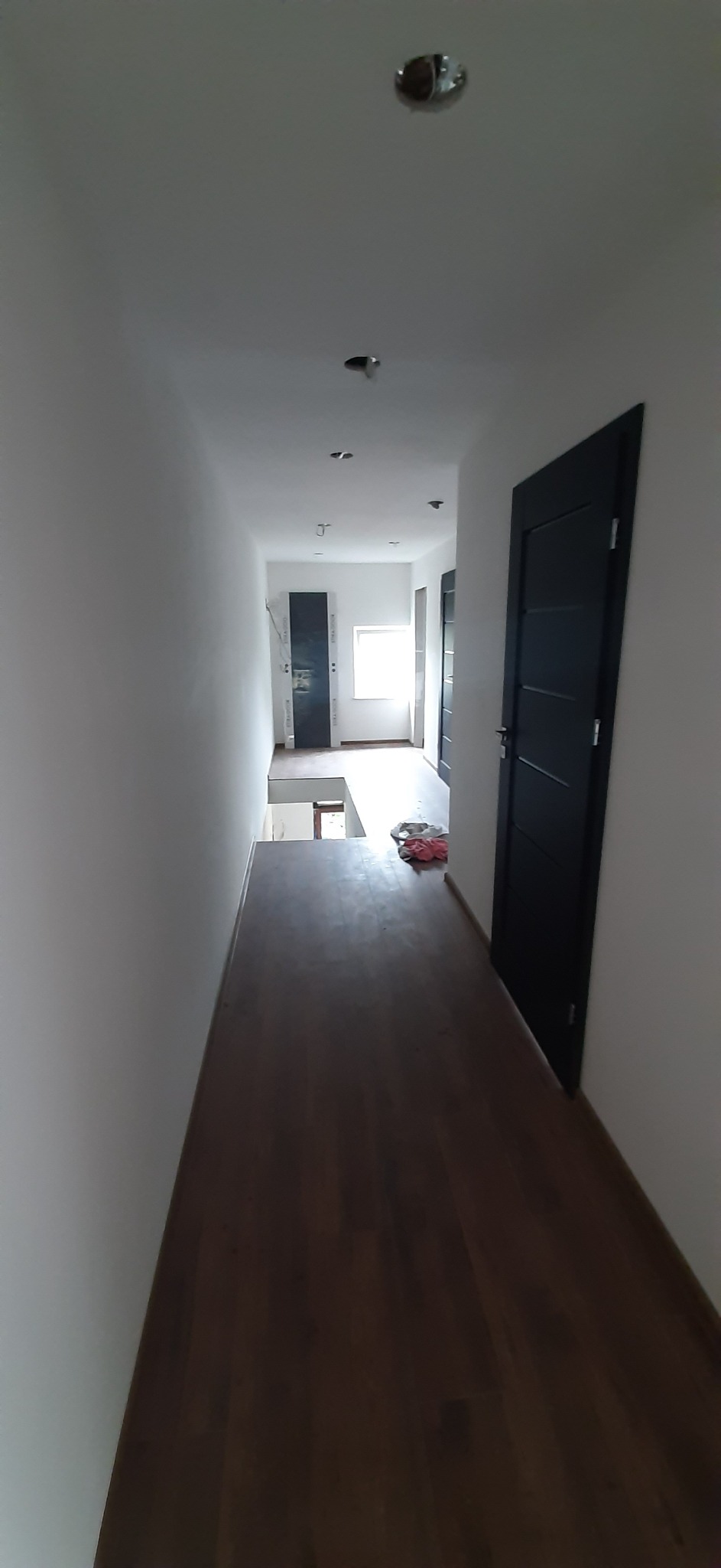 Korytarz z nową, ciemną podłogą z paneli, świeżo pomalowane białe ściany, czarne drzwi po prawej stronie, sufit z zamontowanymi punktami świetlnymi, widok w głąb mieszkania.