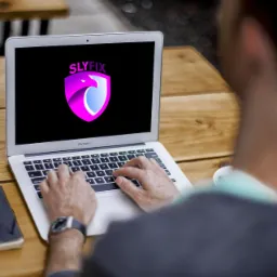 Osoba używa laptopa MacBook Air z wyświetlonym logo Slyfix na ekranie, obok leży iPhone, notatnik i długopis na drewnianym stole.