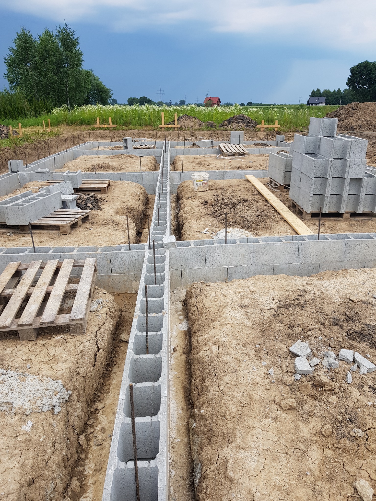 Fundamenty domu w trakcie budowy: bloczki betonowe ułożone w wykopie, widoczne zbrojenia pionowe. W tle łąka i zabudowania.