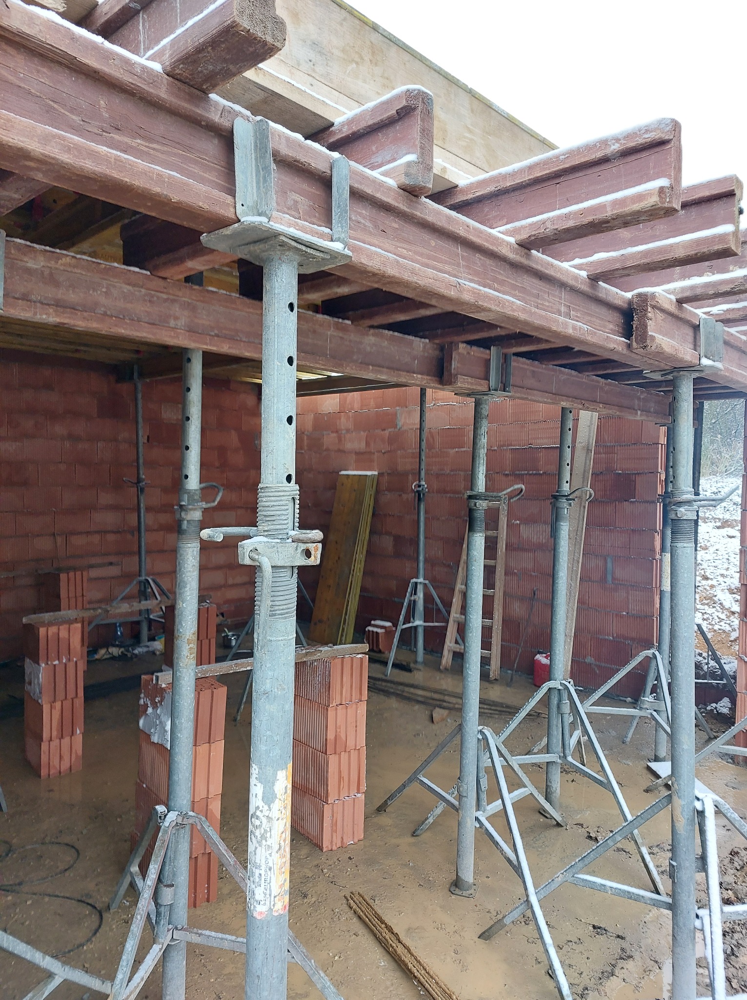 Wznoszenie ścian z czerwonej cegły i szalunku stropu, wspartego na metalowych stemplach. Widoczne bloczki ceglane i drewniane elementy konstrukcyjne. Zimowa aura.