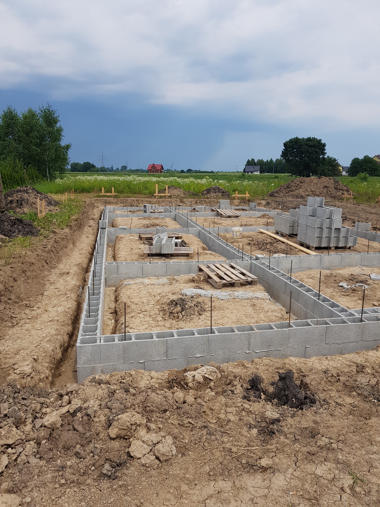 Wykopane fundamenty z bloczków betonowych, zbrojenie pionowe, palety z bloczkami w tle, widok na łąkę i dom w oddali, pochmurne niebo.