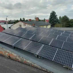 Panele słoneczne zamontowane na płaskim dachu budynku z ceglaną ścianą attyki, widoczne okoliczne domy i pochmurne niebo.