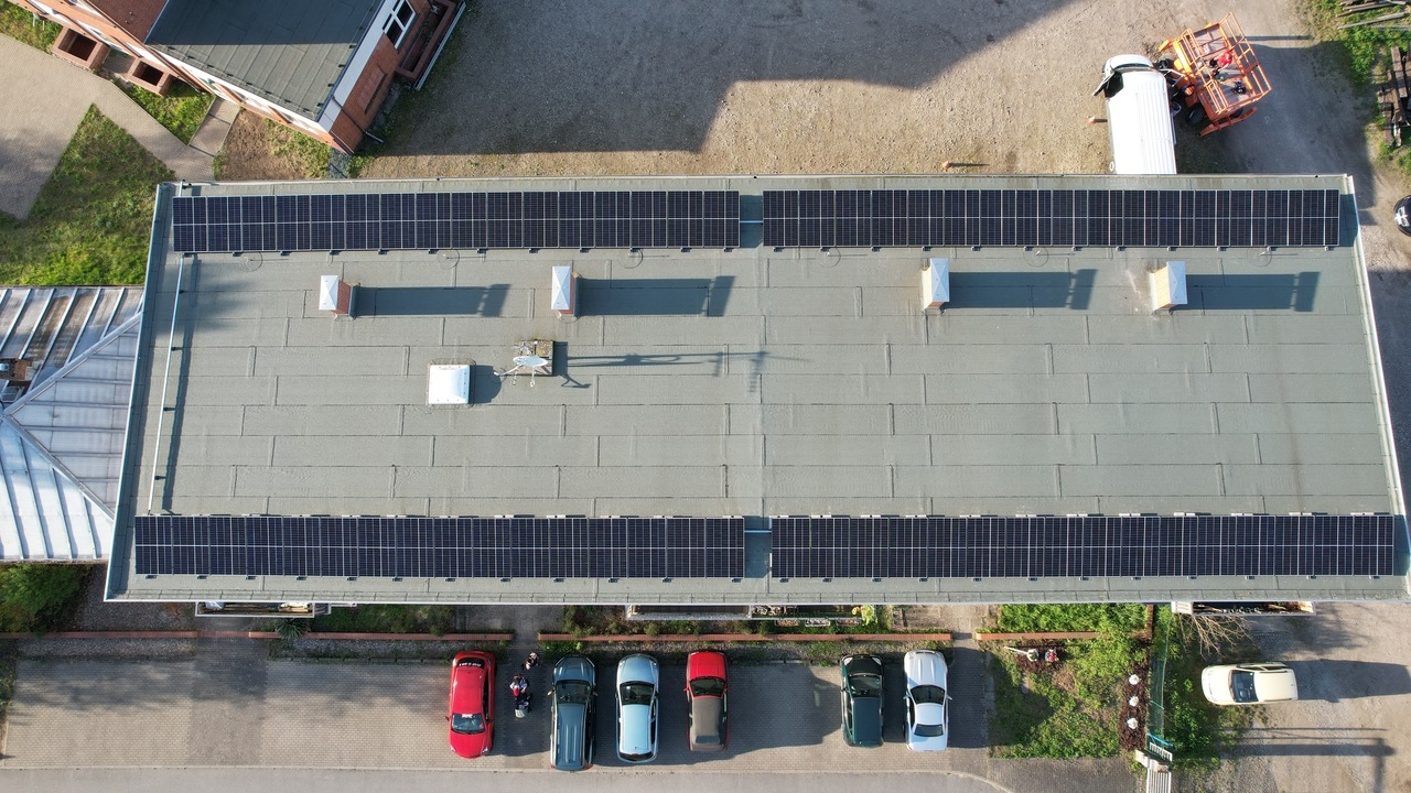 Widok z góry na płaski dach budynku przemysłowego z rzędami paneli słonecznych; w tle widoczny podnośnik koszowy i zaparkowane samochody.