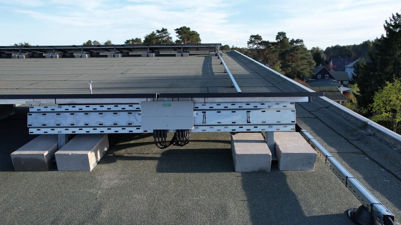 Montaż paneli słonecznych na płaskim dachu z użyciem betonowych bloków jako balastu, widoczna skrzynka przyłączeniowa Projoy PEFS i elementy konstrukcyjne systemu mocowania.
