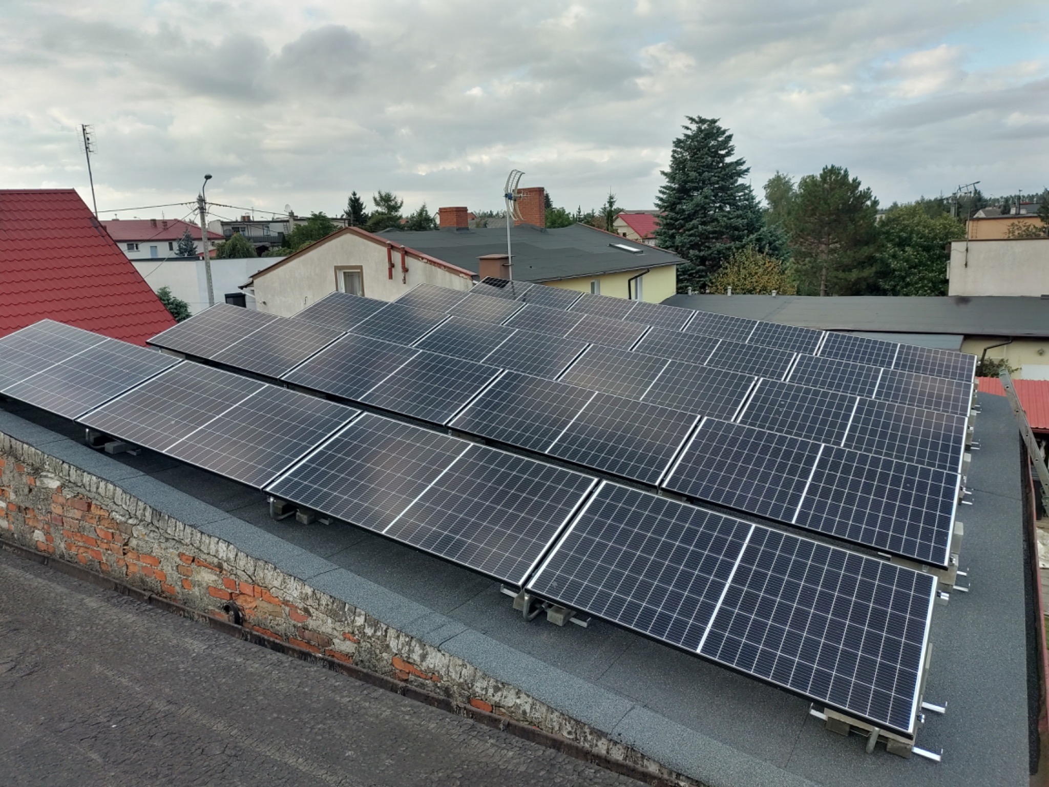 Panele słoneczne zamontowane na płaskim dachu budynku z ceglaną ścianą attyki, widoczne okoliczne domy i pochmurne niebo.