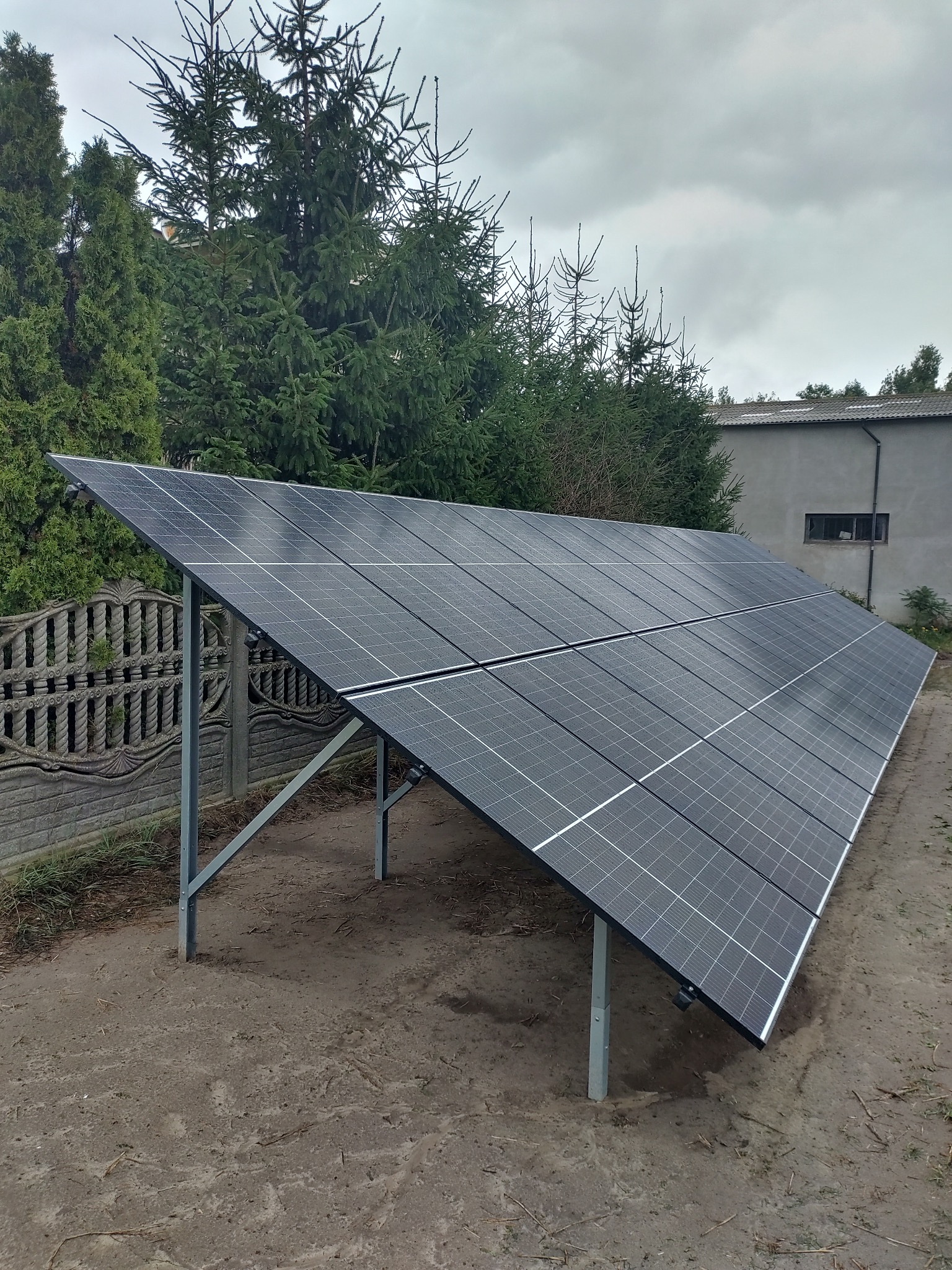Ustawiona na ziemi instalacja paneli słonecznych na metalowej konstrukcji, widok z boku, w tle budynek i drzewa iglaste przy pochmurnym niebie.