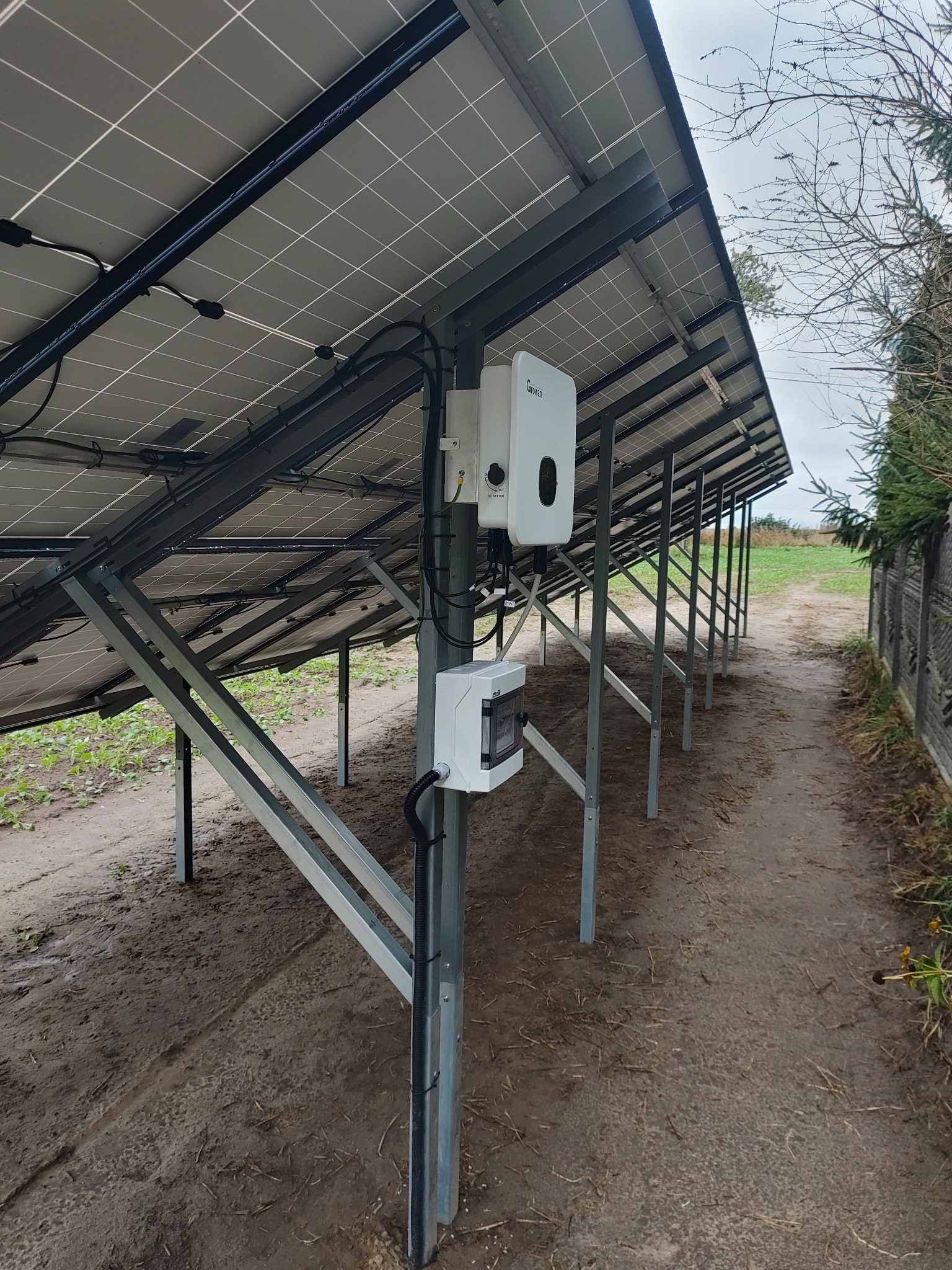Widok od spodu na instalację paneli słonecznych z inwerterem Growatt na metalowej konstrukcji, na zewnątrz, z ziemistym podłożem i fragmentem ogrodzenia.