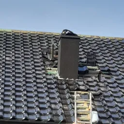 Częściowy widok dachu z czarną dachówką, z fragmentami usuniętej dachówki wokół komina, drabina oparta o dach, nieotynkowana ściana budynku w tle.