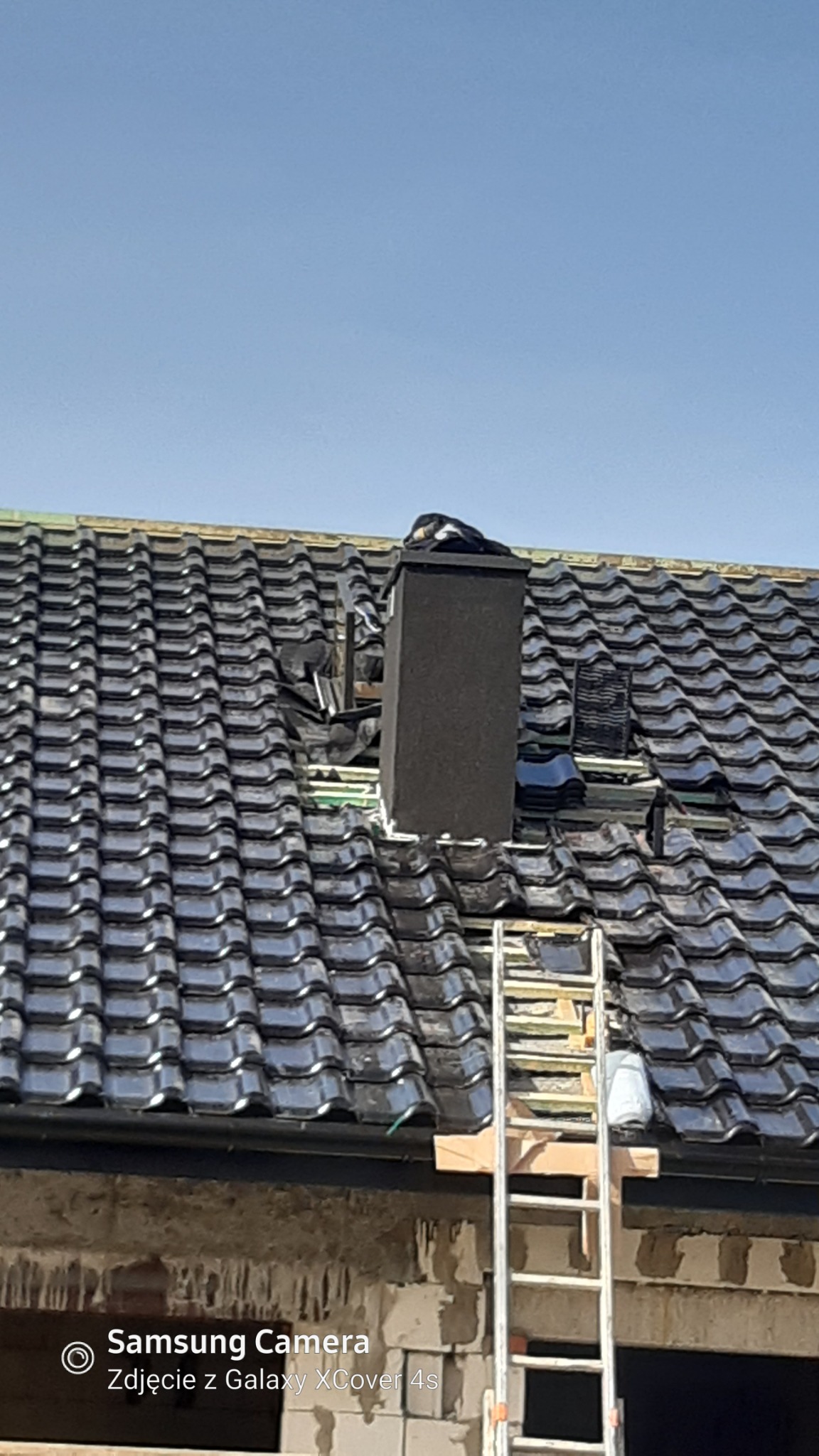 Częściowy widok dachu z czarną dachówką, z fragmentami usuniętej dachówki wokół komina, drabina oparta o dach, nieotynkowana ściana budynku w tle.