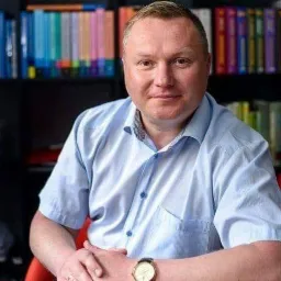 Portret mężczyzny w błękitnej koszuli z zegarkiem na tle regału z książkami o różnej kolorystyce okładek.