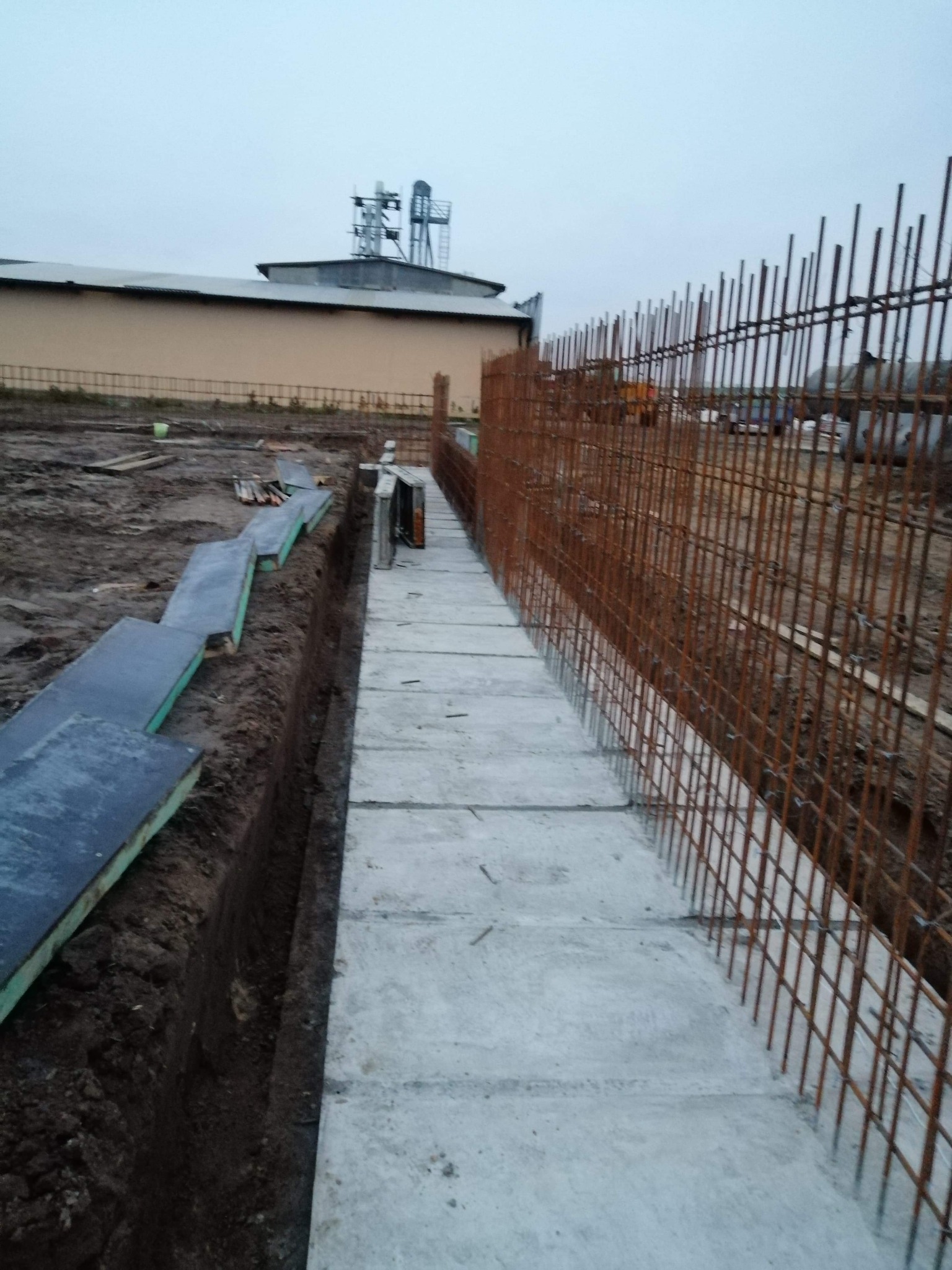 Wykop fundamentowy z ułożonymi betonowymi płytami i zbrojeniem pionowym, widok na budynek gospodarczy w tle.