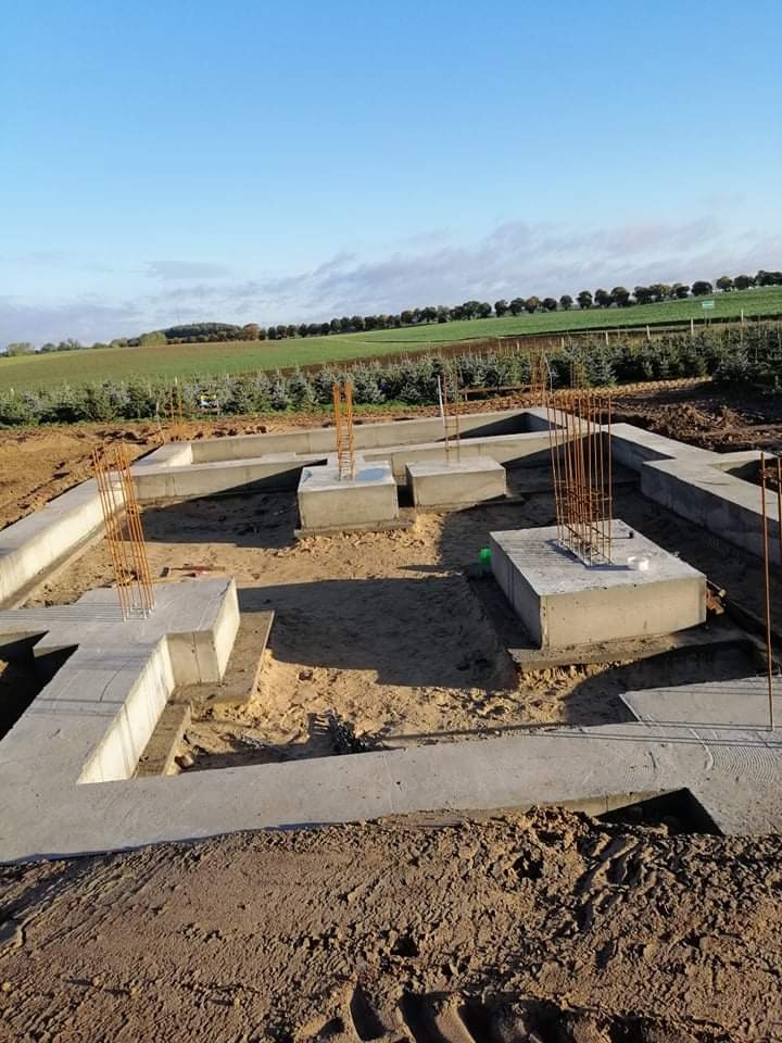 Wylane betonowe fundamenty pod budowę domu z wystającymi prętami zbrojeniowymi, widok na krajobraz z zielonym polem i drzewami w tle.