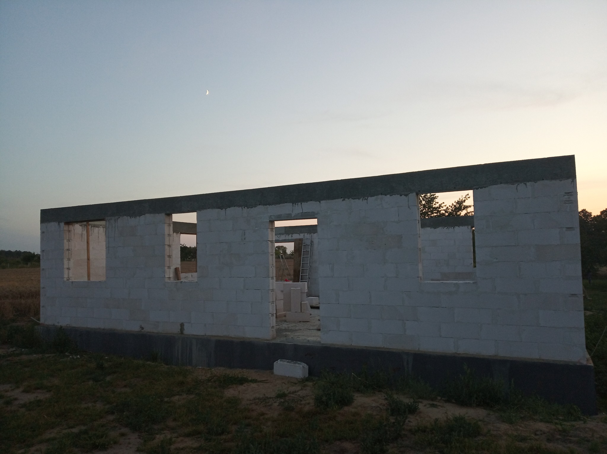 Surowy stan domu z betonu komórkowego w trakcie budowy, widoczne otwory okienne i drzwiowe, na tle wieczornego nieba z cienkim sierpem księżyca.