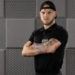 Mężczyzna z brodą i tatuażami na ramionach, ubrany w czarną koszulkę polo z logo 'Reha Fit', stoi na tle szarej ściany z geometrycznym wzorem.