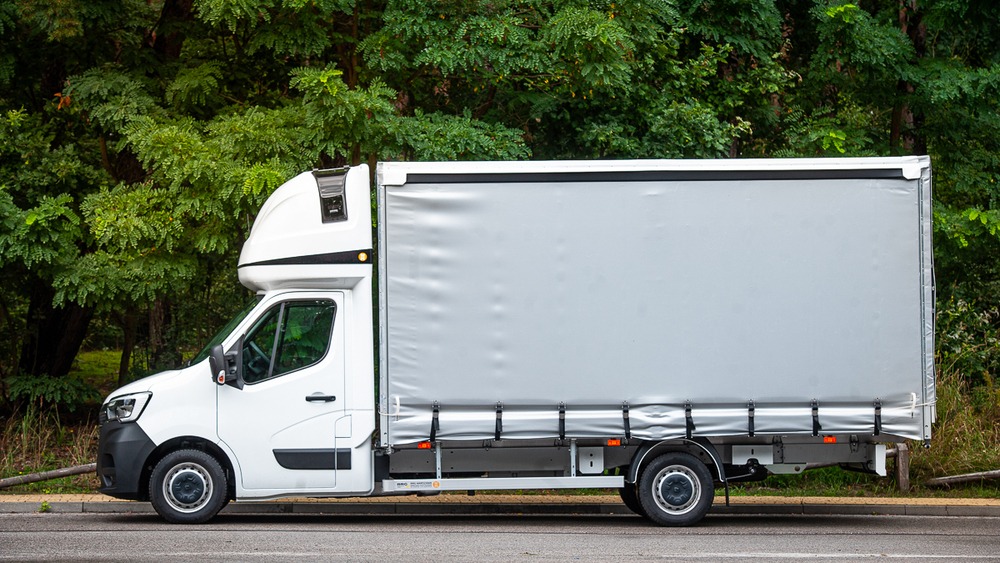Biały bus dostawczy z plandeką na tle zieleni, zaparkowany na poboczu, widok z boku.