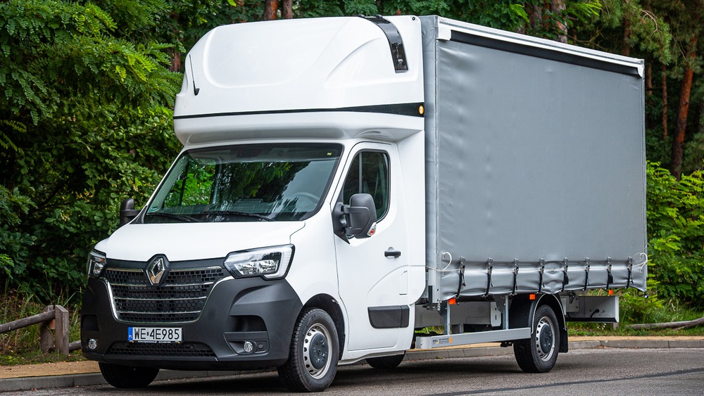 Biały samochód dostawczy Renault Master z plandeką i wysoką kabiną, zaparkowany na tle zieleni.