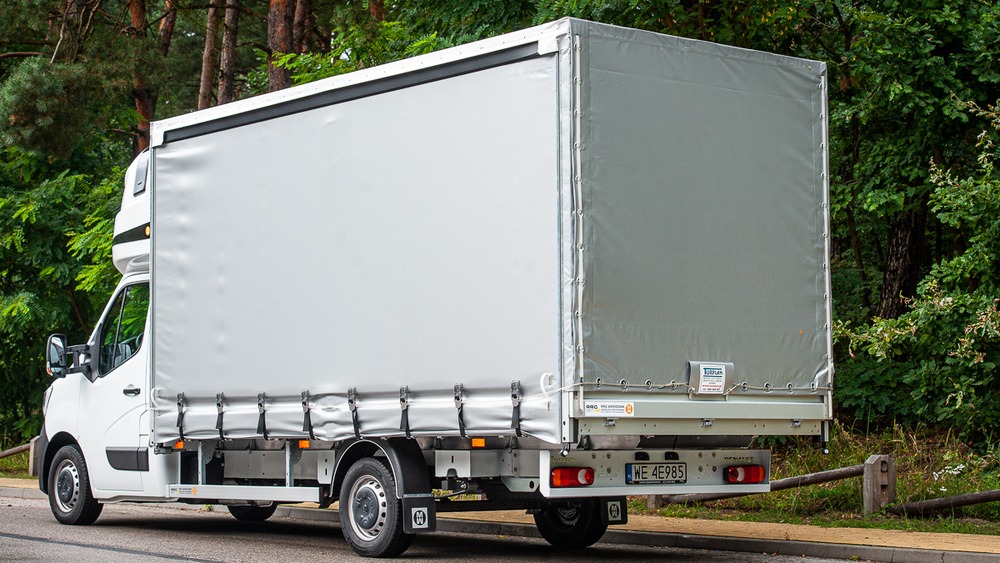 Biały samochód dostawczy z plandeką, zaparkowany na asfalcie, widok z tyłu na tle lasu, tablica rejestracyjna WE 4E985.