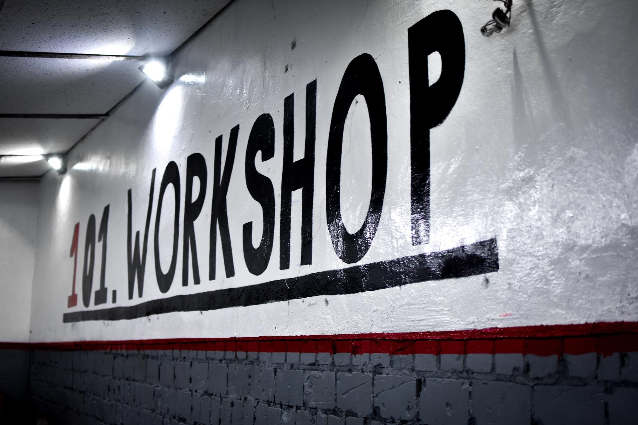 Malowany napis '101. WORKSHOP' na ścianie warsztatu, widok z dołu, oświetlenie punktowe.