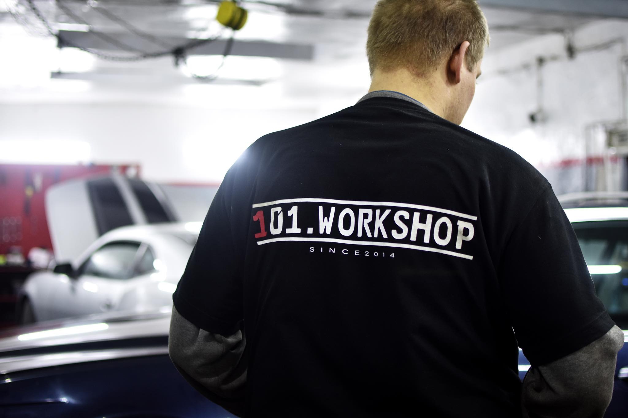 Mężczyzna tyłem w czarnej koszulce z białym napisem '101.WORKSHOP SINCE 2014' na tle warsztatu samochodowego z dwoma autami, w tym jednym z otwartą maską.