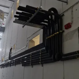 Profesjonalnie poprowadzone, zaizolowane rury instalacji klimatyzacyjnej wzdłuż szarej, betonowej ściany wewnątrz budynku. Widoczne uchwyty mocujące i elementy wentylacji.